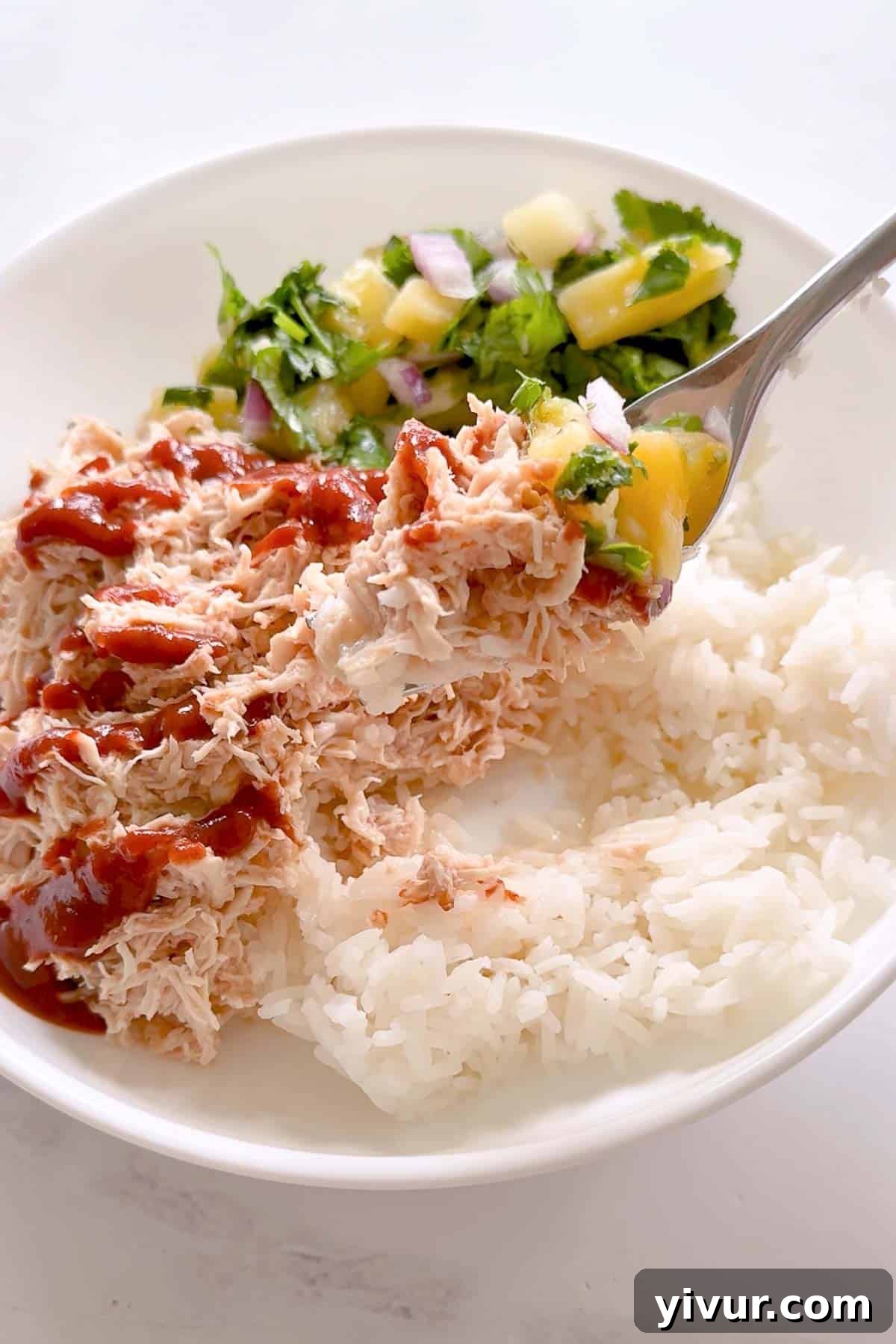 A beautifully presented white bowl featuring High Protein Instant Pot Hawaiian Chicken Bowls. Shredded, saucy chicken is piled on a bed of white rice, generously topped with extra barbecue sauce and a vibrant pineapple salsa. The salsa, a mix of cilantro, red onion, and pineapple chunks, adds a fresh, zesty counterpoint. A fork, holding some salsa, hovers above the inviting meal.