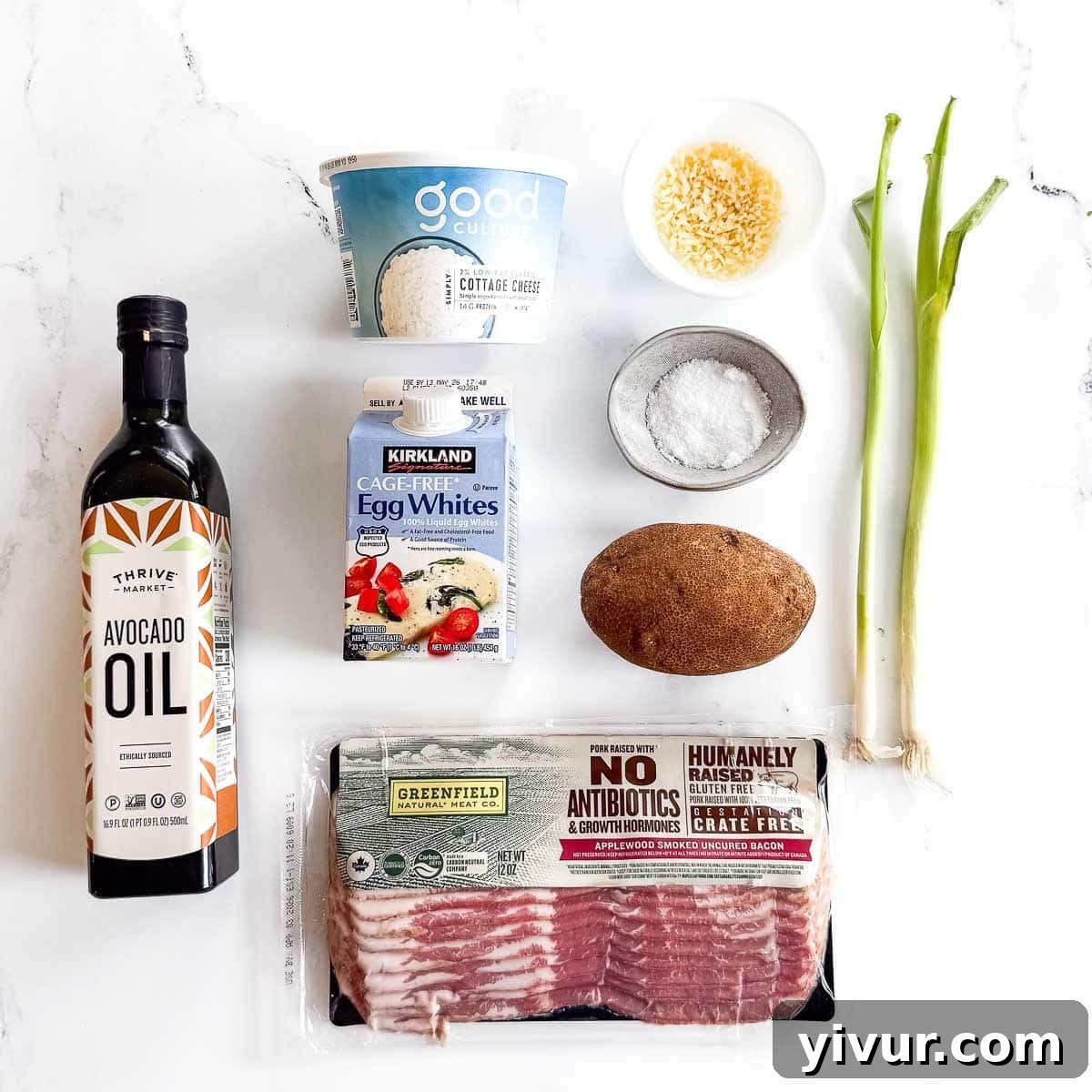 A flat lay of high protein breakfast ingredients on a white surface, including avocado oil, cottage cheese, egg whites, grated cheese, salt, a russet potato, green onions, and uncooked bacon.
